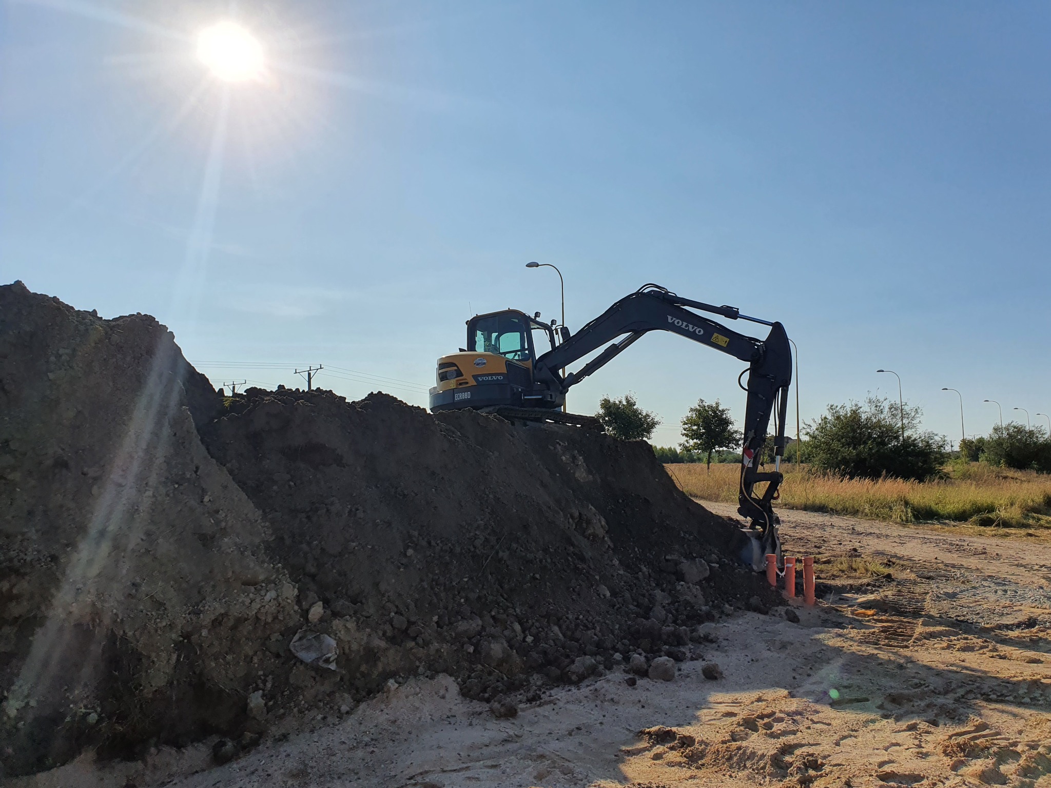 Żółto-czarna koparka Volvo na placu budowy, ustawiona na wzniesieniu ziemi, z trzema pomarańczowymi rurami wystającymi z ziemi, oświetlona jasnym słońcem.