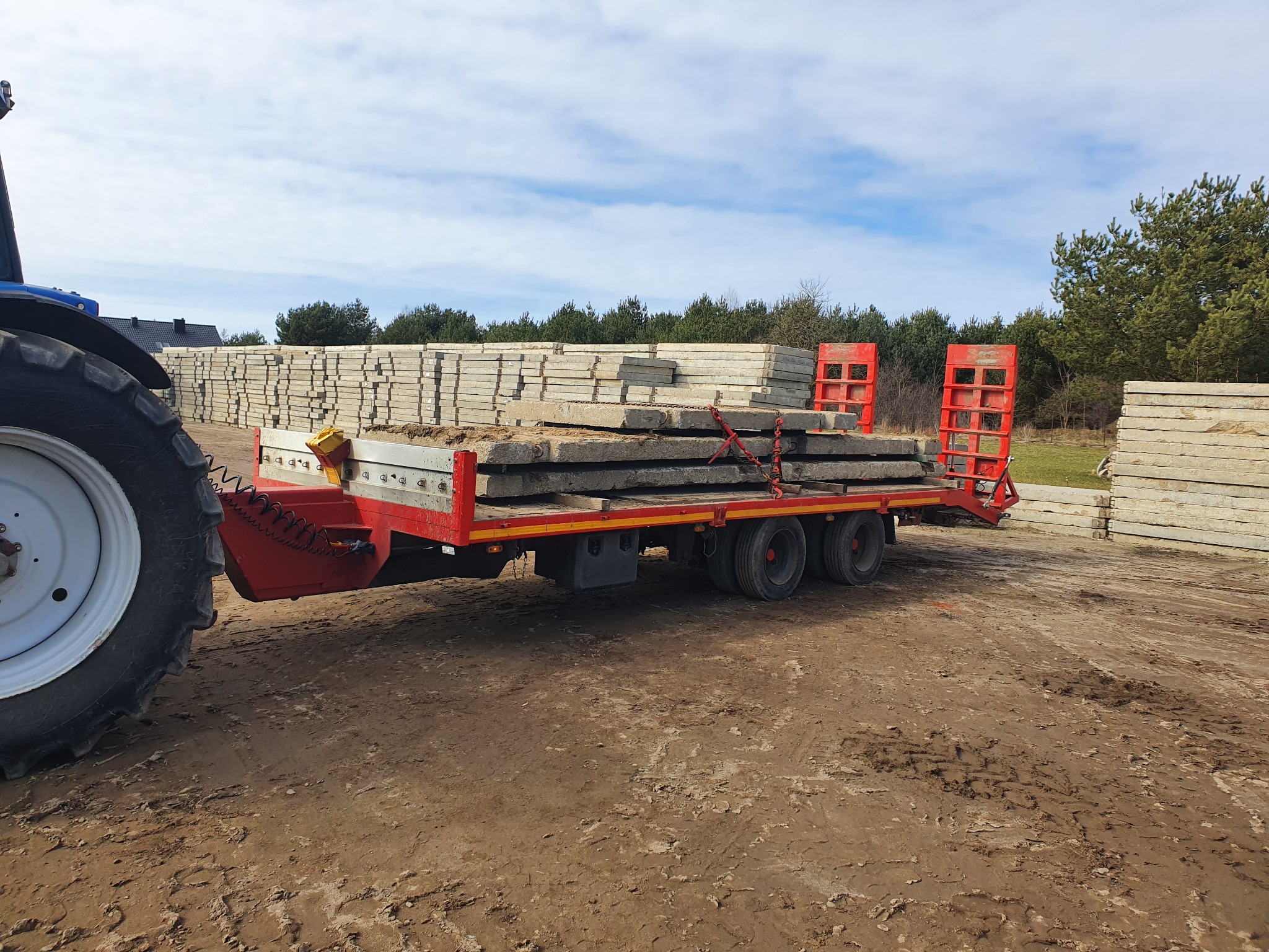 Czerwona przyczepa z ładunkiem betonowych płyt, ciągnięta przez traktor na błotnistym placu budowy, w tle stosy płyt i drzewa.