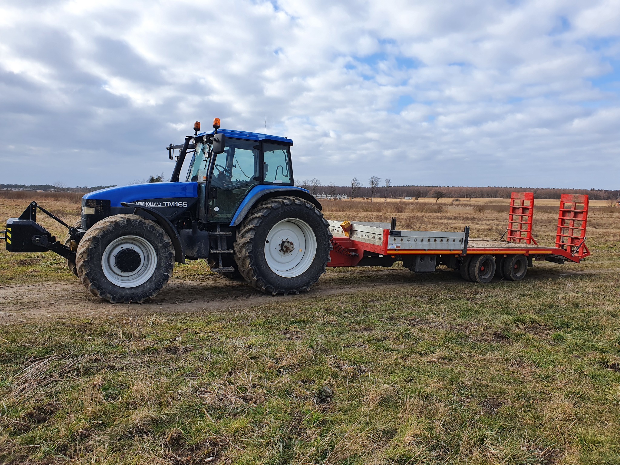 Niebieski traktor New Holland TM165 z czerwoną przyczepą platformową na polu z suchą trawą, pochmurne niebo w tle.
