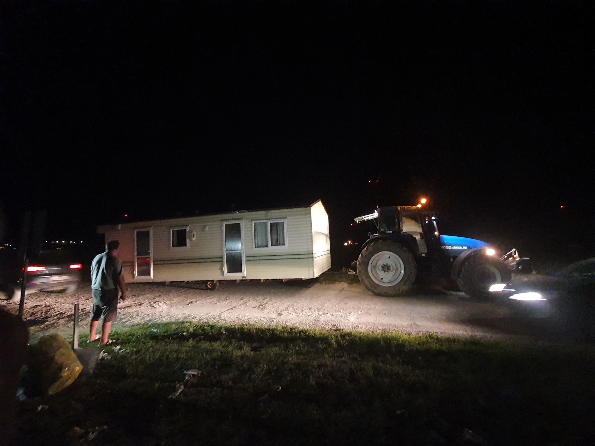 Ciężki traktor New Holland holuje mobilny domek letniskowy w nocy; widoczna sylwetka mężczyzny i światła samochodu w tle.
