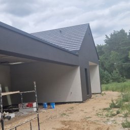 Zakład Ogólnobudowlany - Dwukolorowa elewacja domu z szarą dachówką, widok z zewnątrz, rusztowanie z boku, teren budowy, las w tle, pochmurne niebo.