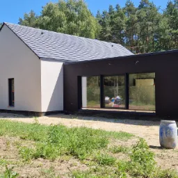 Nowoczesny dom jednorodzinny z elewacją w dwóch kolorach: jasnym i ciemnym, z dużymi oknami i szarym dachem, otoczony zielenią i drzewami.