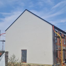 Zakład Ogólnobudowlany - Świeżo otynkowana, biała ściana domu jednorodzinnego z widocznymi rusztowaniami i fragmentem elewacji w stanie surowym. Okno zabezpieczone folią.