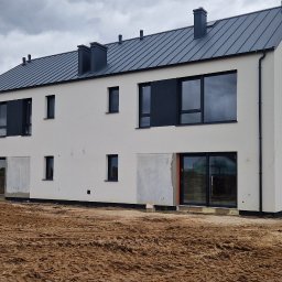 Zakład Ogólnobudowlany - Dwukondygnacyjny dom w zabudowie bliźniaczej z szarą blachodachówką i białymi ścianami, częściowo otynkowany, z widocznym fragmentem busa. Pochmurne niebo.