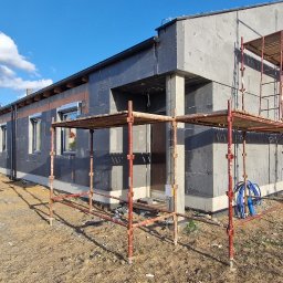 Zakład Ogólnobudowlany - Dom jednorodzinny w trakcie ocieplania, widoczne rusztowania i czarny styropian. Częściowo ocieplona elewacja, błękitne niebo w tle. Na pierwszym planie cień fotografa.