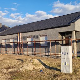 Zakład Ogólnobudowlany - Dom w trakcie ocieplania, widoczne rusztowania i styropian na elewacji. Okna zabezpieczone folią. Na pierwszym planie skrzynka elektryczna.