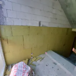 Wnętrze poddasza w trakcie ocieplania wełną mineralną, widoczne bloki betonowe powyżej izolacji, fragment okna po lewej stronie i opakowanie wełny na podłodze.