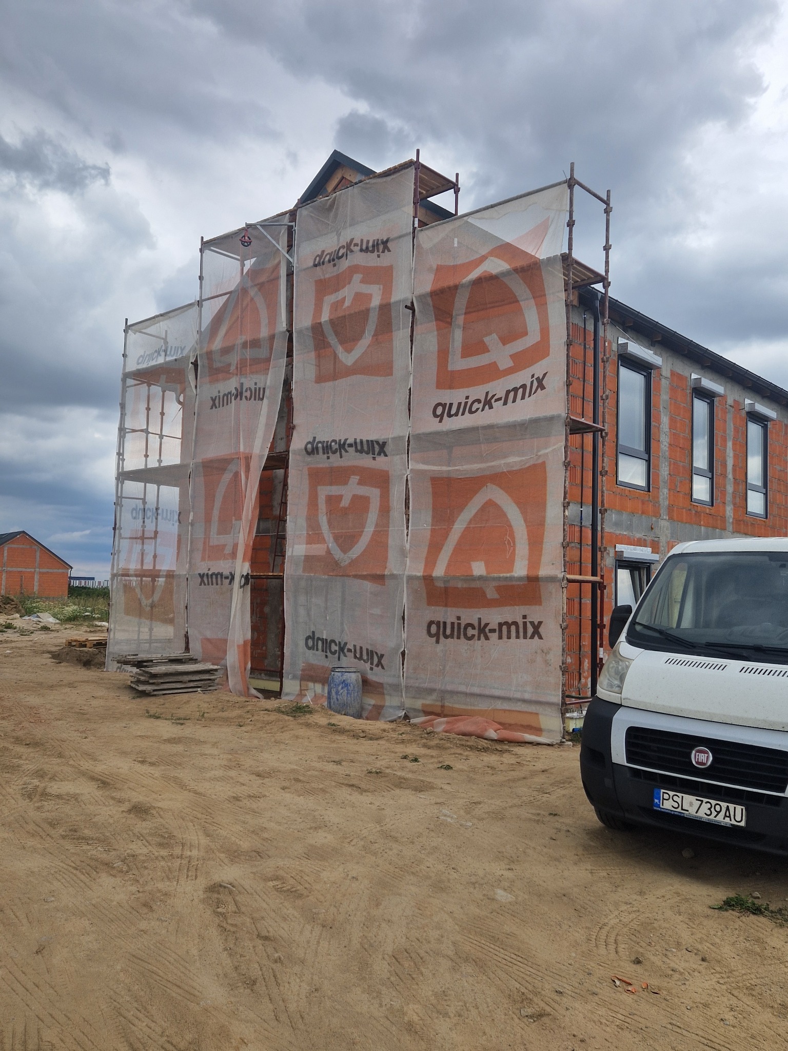 Dom w trakcie budowy, osłonięty siatką z logo Quick-Mix na rusztowaniu. Widoczne cegły i okna, obok zaparkowany biały bus Fiat. Pochmurne niebo w tle.