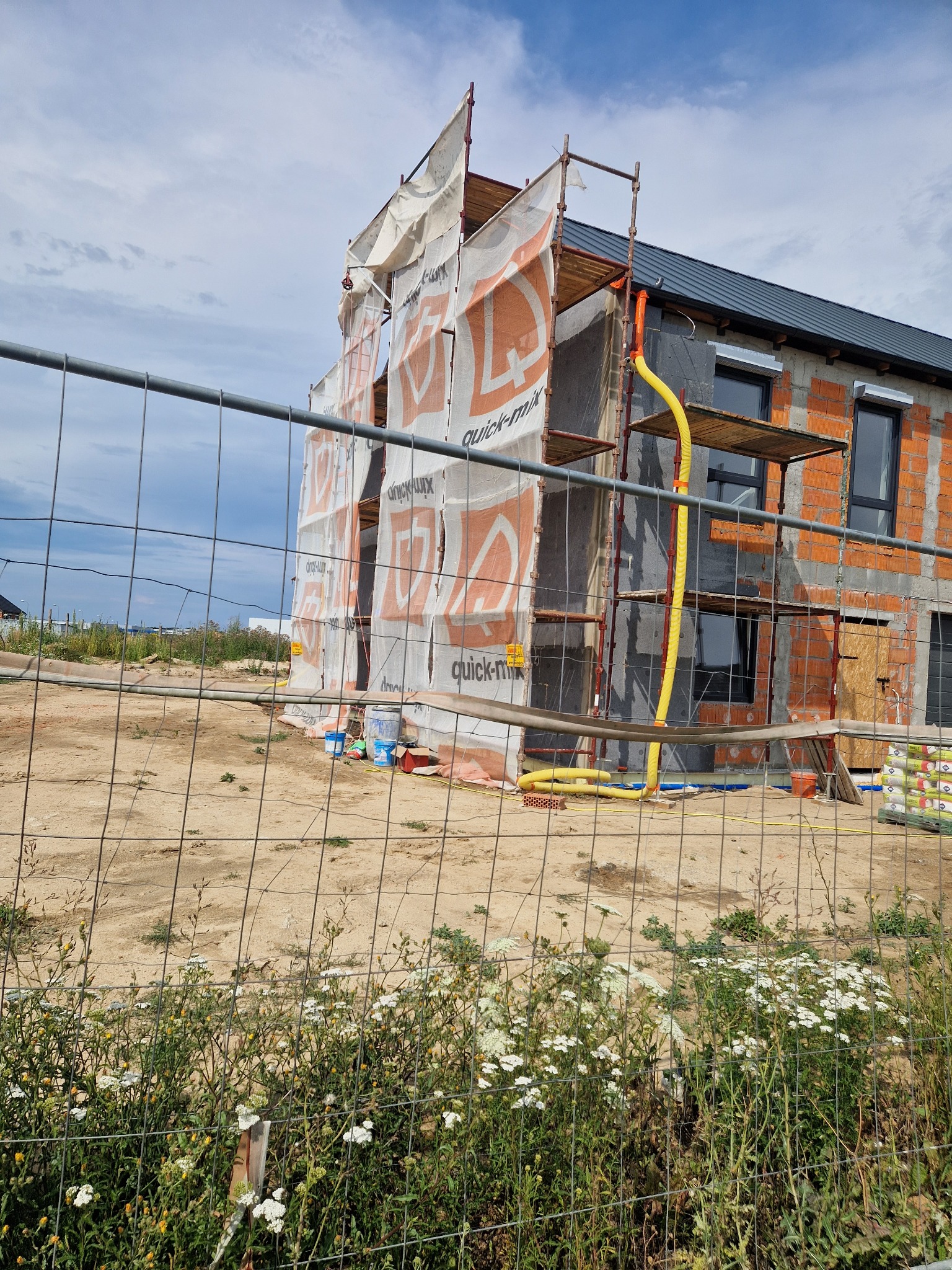 Budowa domu w trakcie ocieplania. Widoczne rusztowania, materiały izolacyjne i częściowo otynkowana ściana z oknami. Żółte rury wentylacyjne. Całość widziana przez metalowe ogrodzenie.