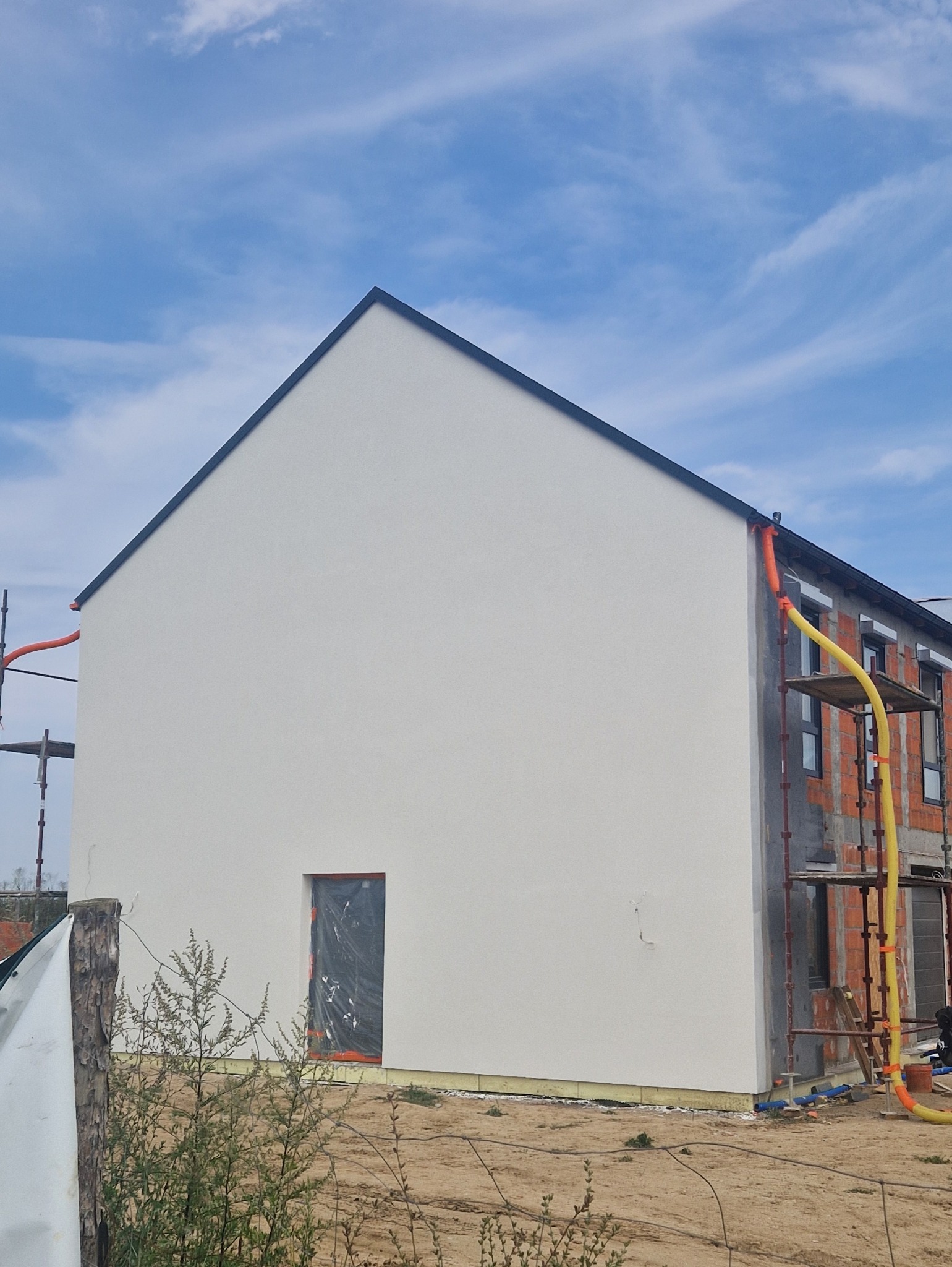 Świeżo otynkowana, biała ściana domu jednorodzinnego z widocznymi rusztowaniami i fragmentem elewacji w stanie surowym. Okno zabezpieczone folią.