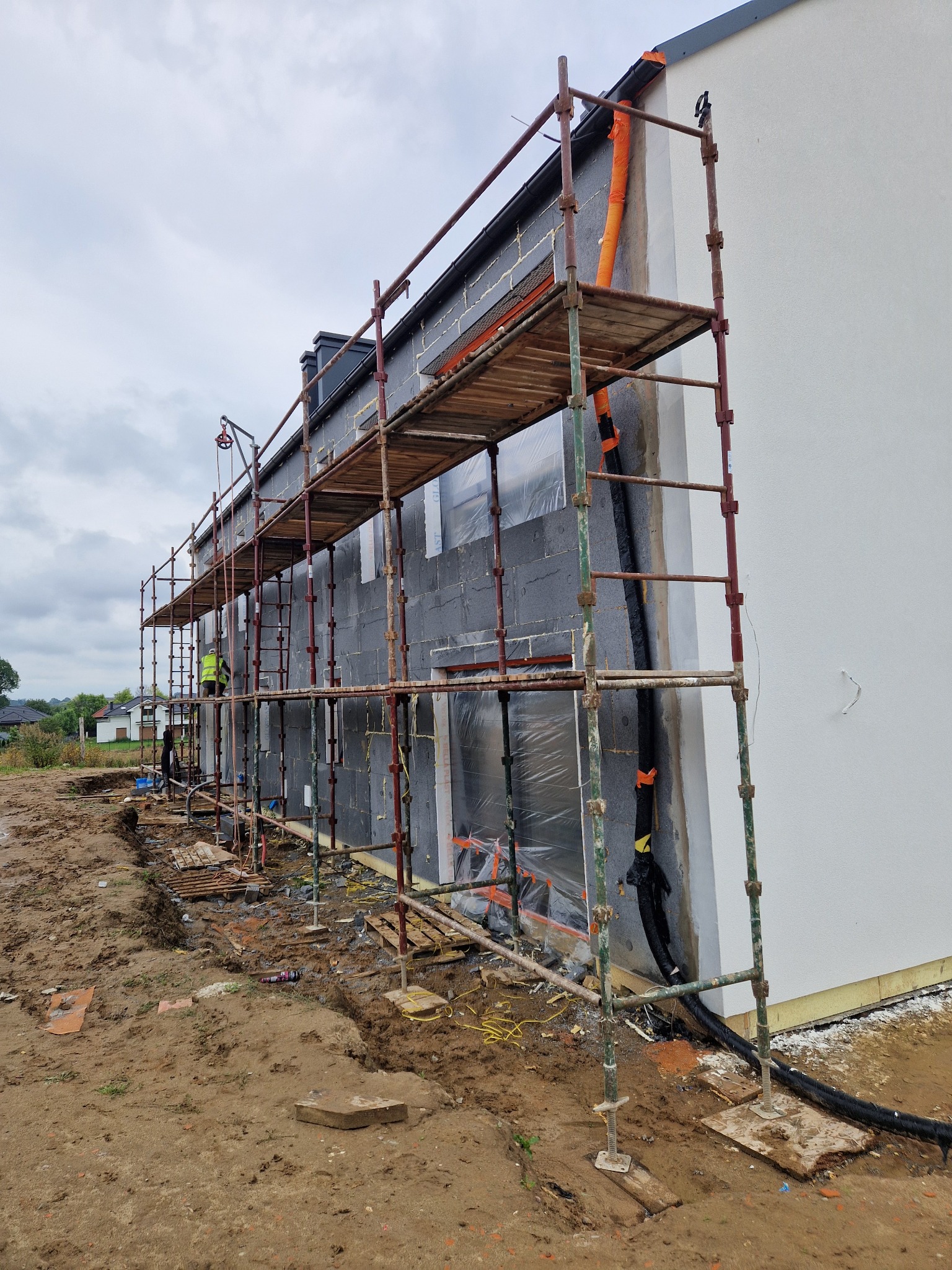 Dom w trakcie ocieplania: szare bloczki, rusztowanie, pracownik w kamizelce. Część elewacji otynkowana na biało. Widoczne instalacje i teren budowy.