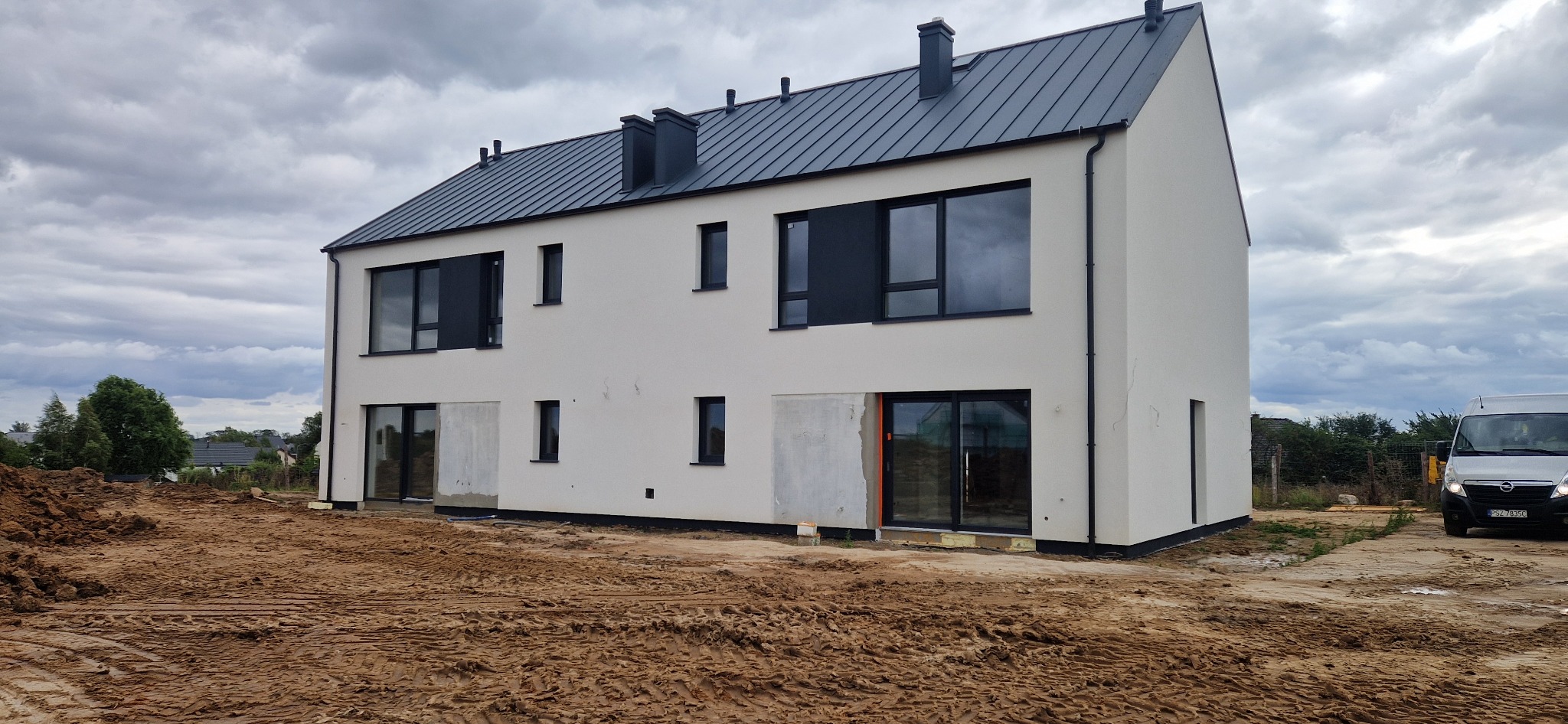 Dwukondygnacyjny dom w zabudowie bliźniaczej z szarą blachodachówką i białymi ścianami, częściowo otynkowany, z widocznym fragmentem busa. Pochmurne niebo.