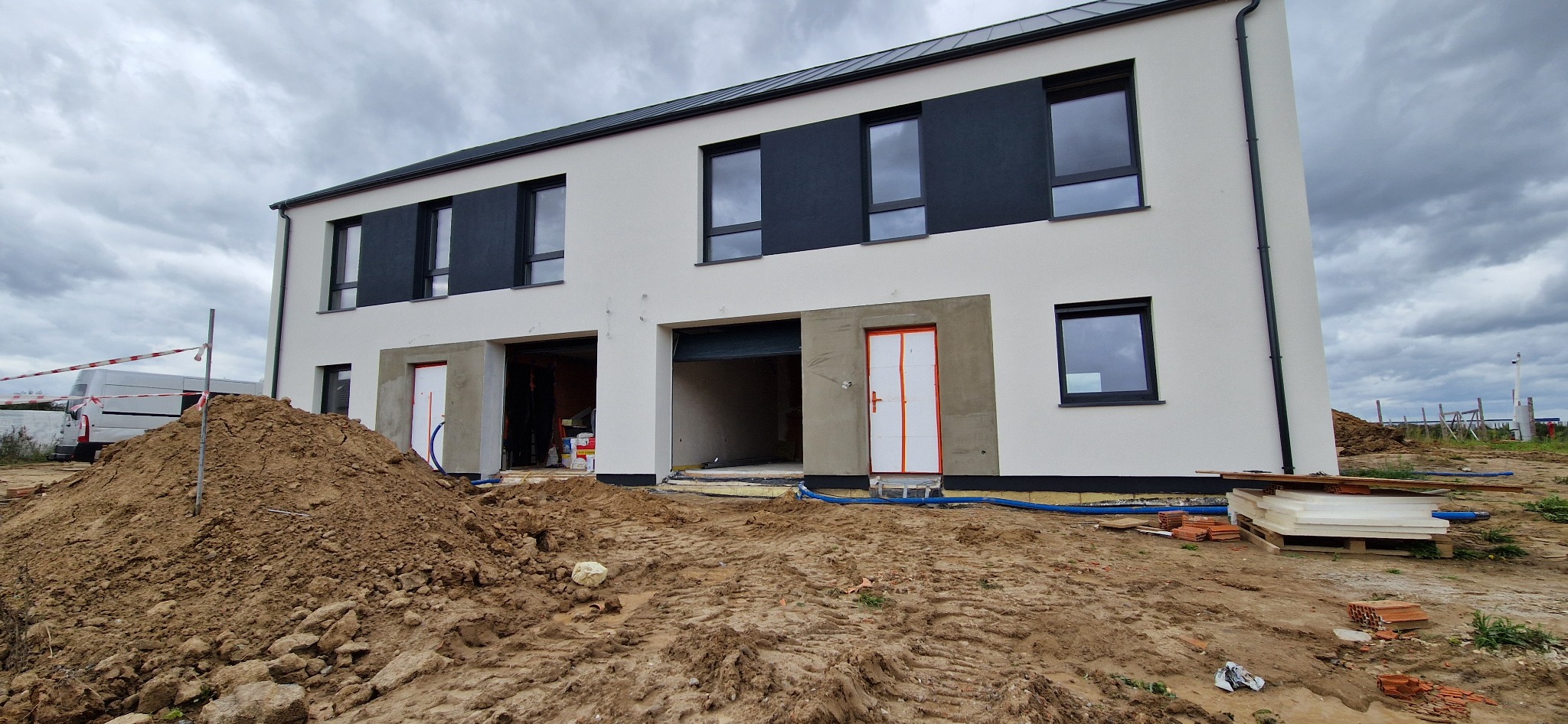 Dwukondygnacyjny budynek w trakcie prac elewacyjnych: fragmenty otynkowane, widoczne materiały budowlane i hałda ziemi na pierwszym planie. Szara, pochmurna pogoda.