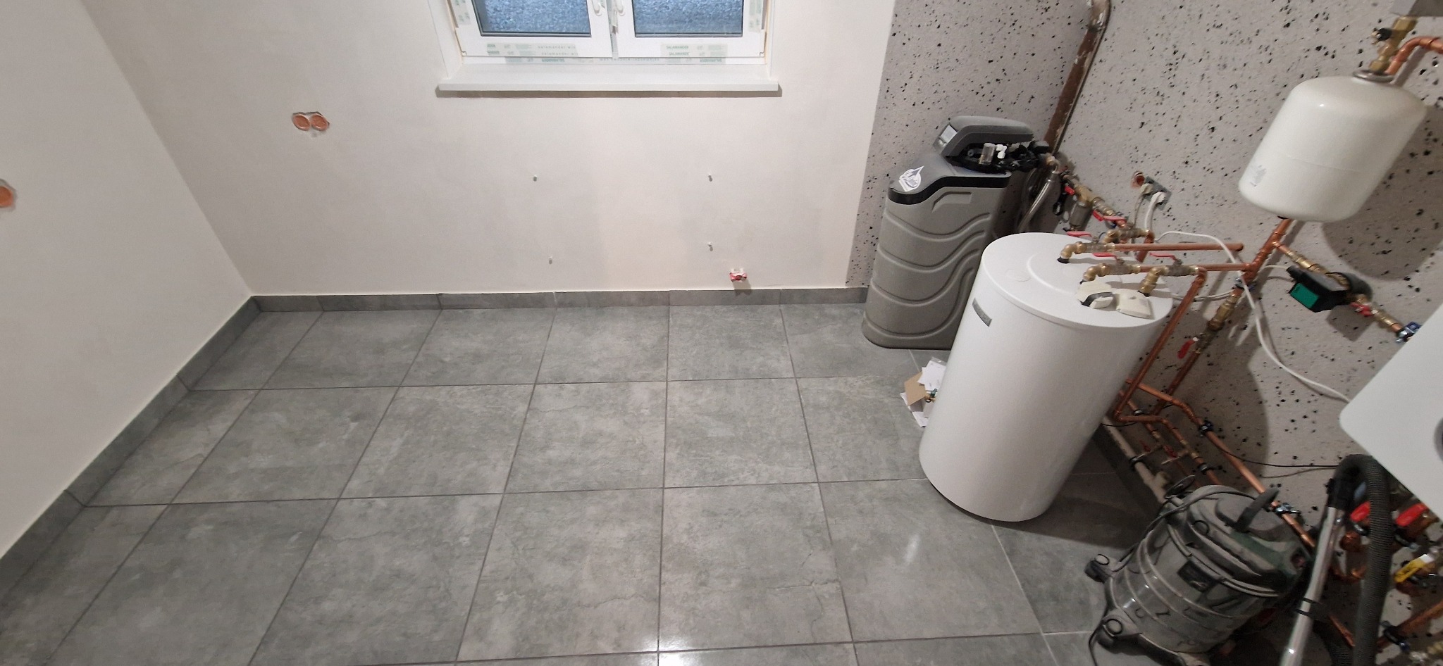 Szare płytki podłogowe w pomieszczeniu z instalacją wodną i bojlerem w Dusznikach. Widoczne przyłącza miedziane i okno w tle. Ściany pomalowane na jasny kolor.