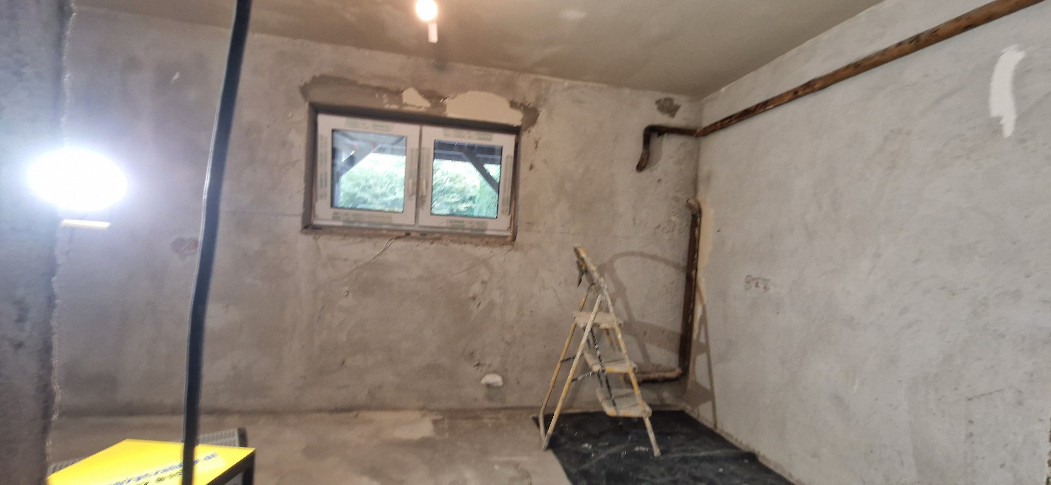 Wnętrze w trakcie prac wykończeniowych: surowe ściany, okno z widokiem na zieleń, drabina malarska i lampa budowlana. Przygotowanie do tynkowania lub szpachlowania.
