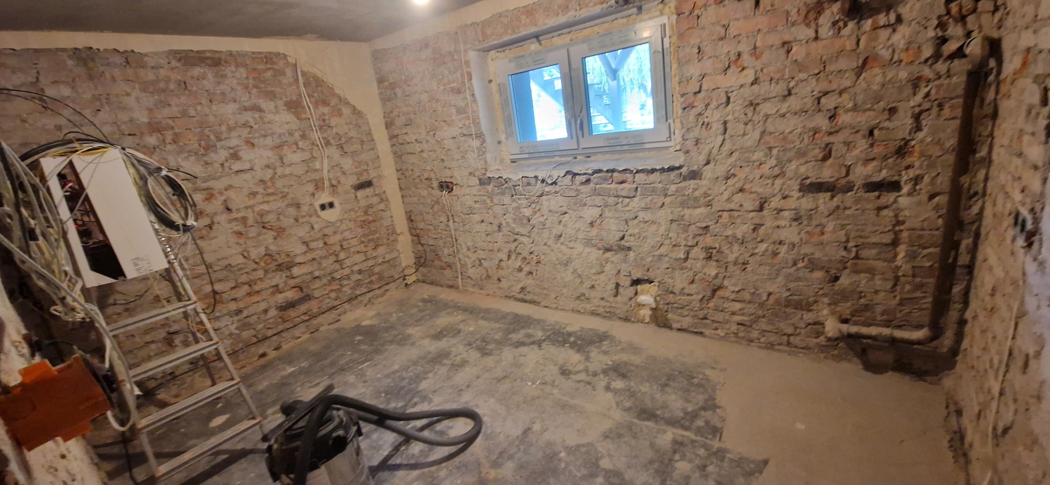 Surowe wnętrze z ceglanymi ścianami przed remontem. Widoczne przewody, drabina i odkurzacz. Okno z widokiem na zewnątrz. Fragment ściany z nałożonym tynkiem.