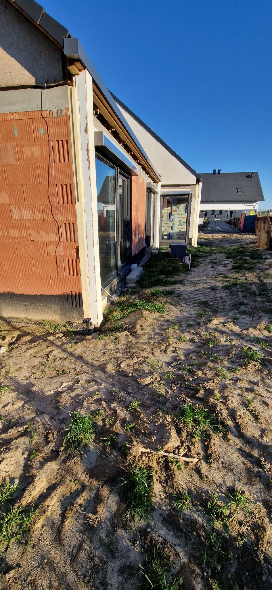 Dom w trakcie ocieplania: widoczne bloczki, izolacja termiczna wokół okien i surowy teren. Prace budowlane w toku, w tle inny budynek.