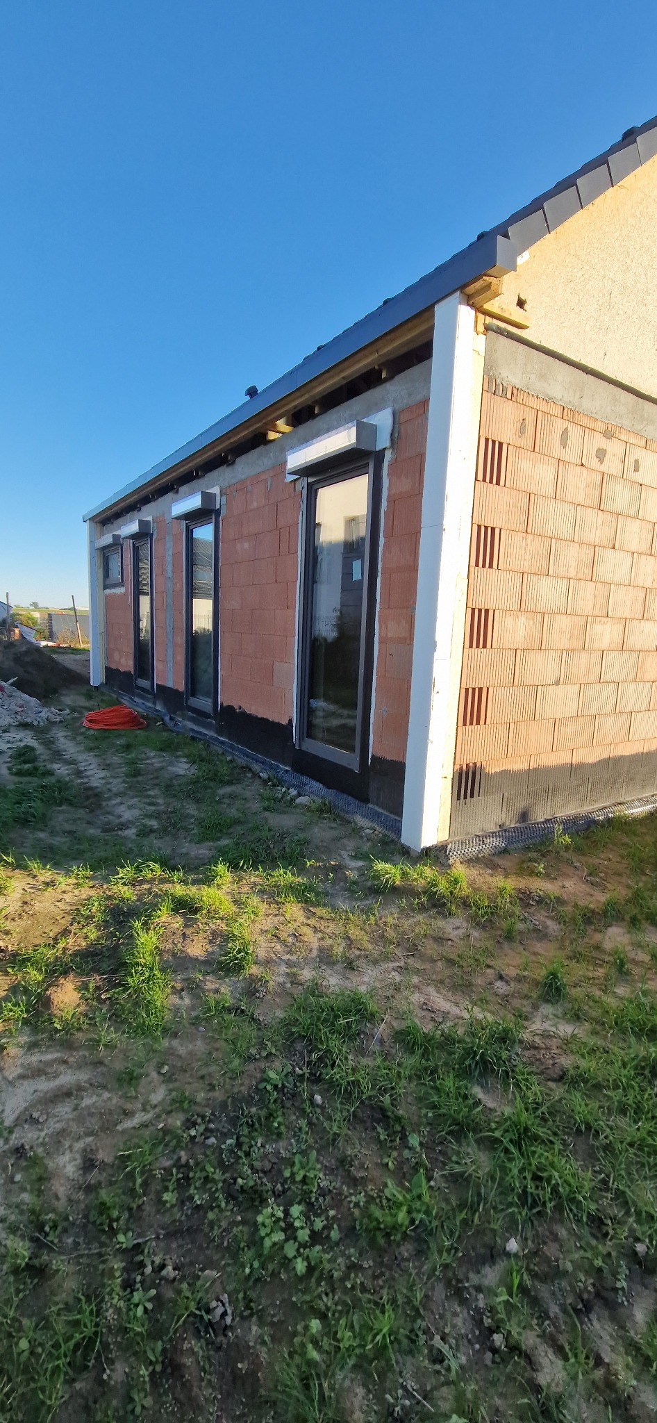 Niedokończony budynek z cegły z oknami i częściowo ocieploną elewacją, widok z zewnątrz na tle błękitnego nieba, w słoneczny dzień.