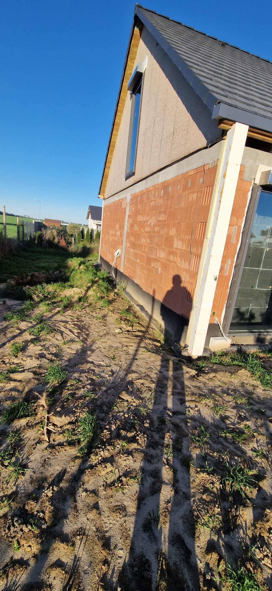 Ściana domu z pustaków, częściowo ocieplona styropianem, z widocznym oknem i fragmentem dachu. Na pierwszym planie cień osoby na ziemi.