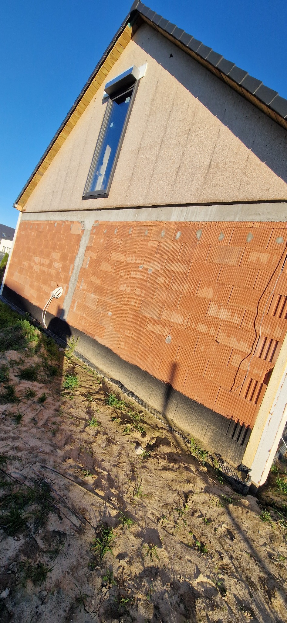 Ściana budynku w trakcie budowy: fragment z cegły ceramicznej, okno dachowe. Widoczne ocieplenie na górnej części ściany oraz izolacja przeciwwilgociowa u dołu.