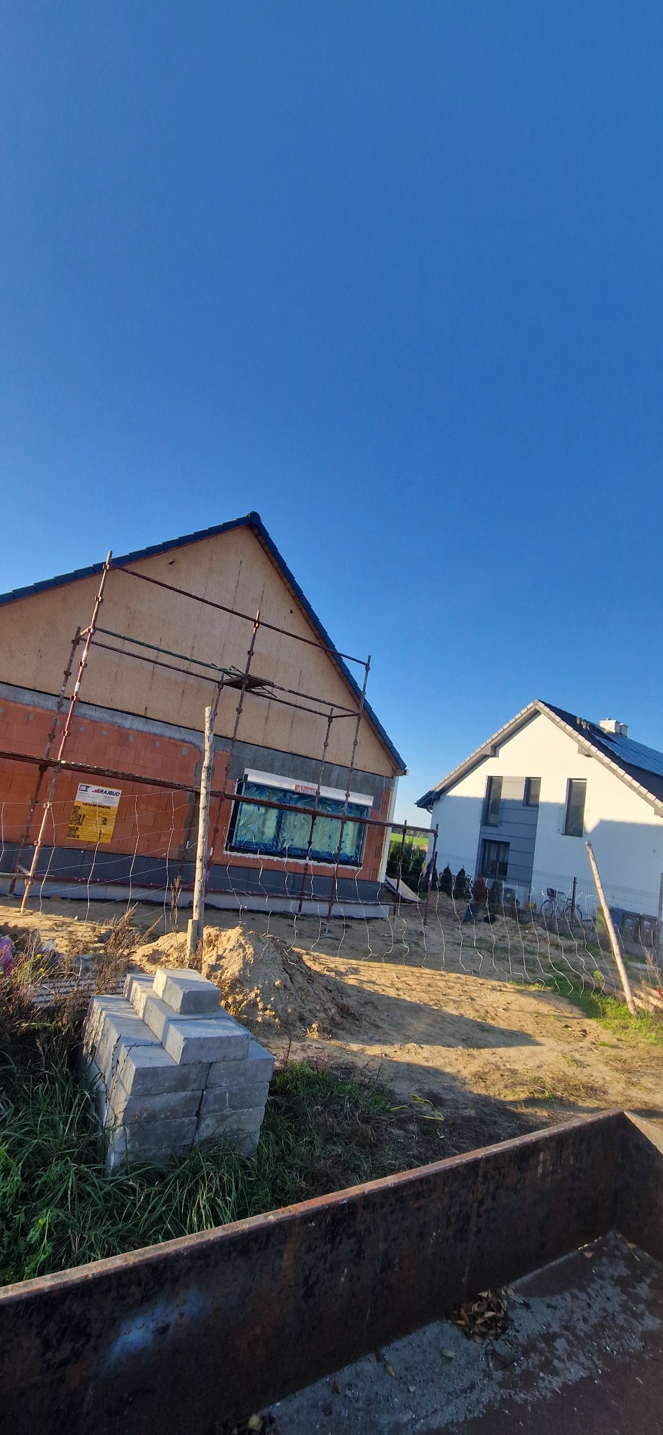 Dom w trakcie budowy, z widocznym rusztowaniem, oknami zabezpieczonymi folią i częściowo otynkowaną elewacją. Obok stoi drugi, zamieszkały budynek.