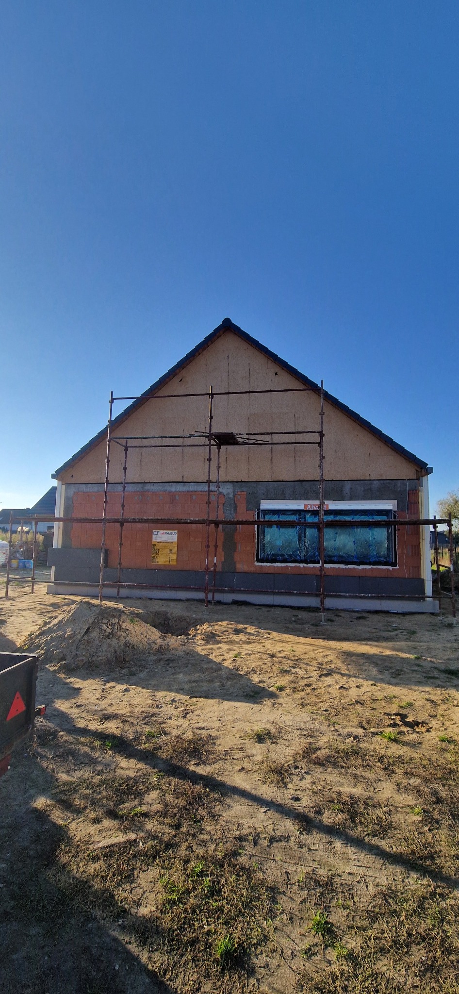 Dom w trakcie ocieplania: widoczne rusztowania, cegły, izolacja i okno zabezpieczone folią. Prace budowlane w toku w słoneczny dzień.