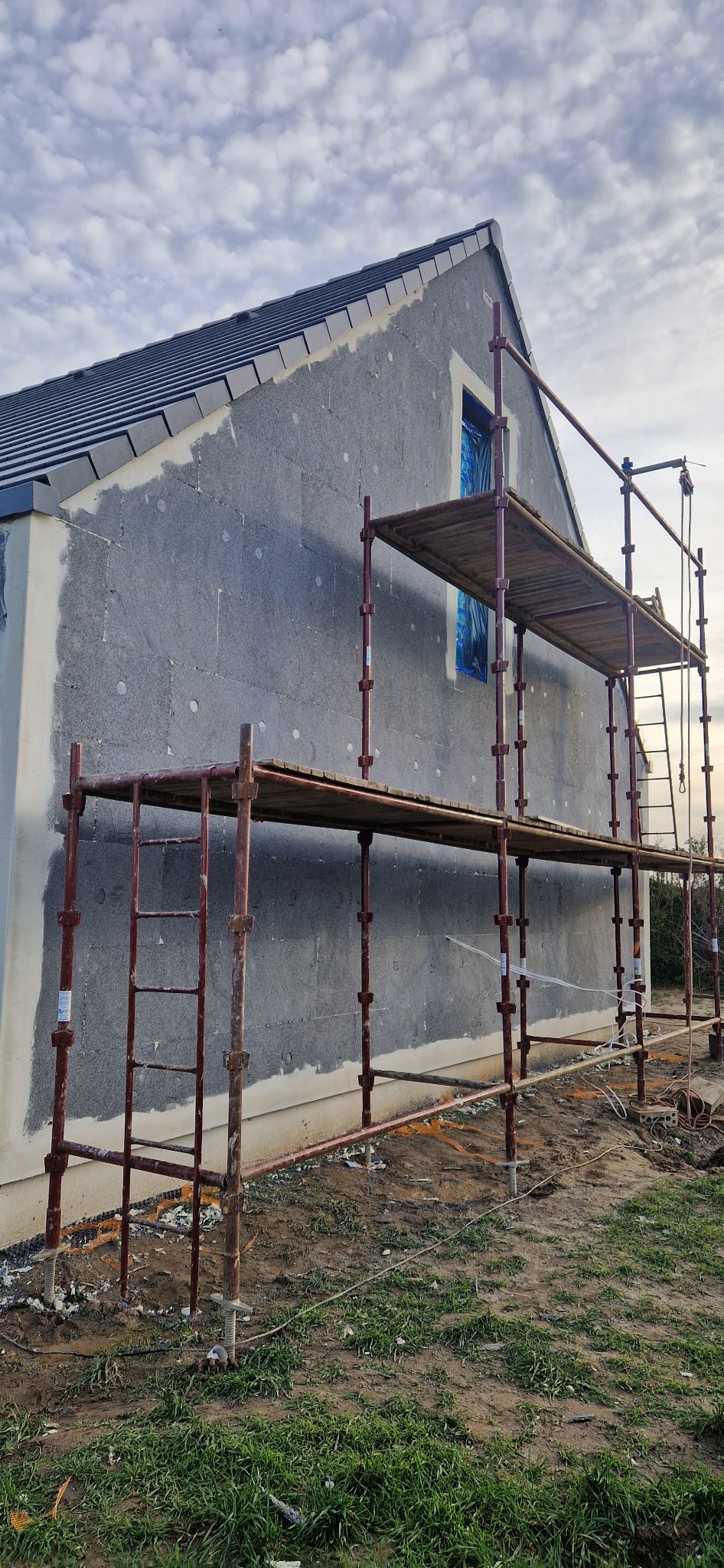Dom w trakcie ocieplania: szara elewacja z widocznym styropianem, zabezpieczone okno folią, rusztowanie. Częściowo otynkowana ściana boczna. Pochmurne niebo.