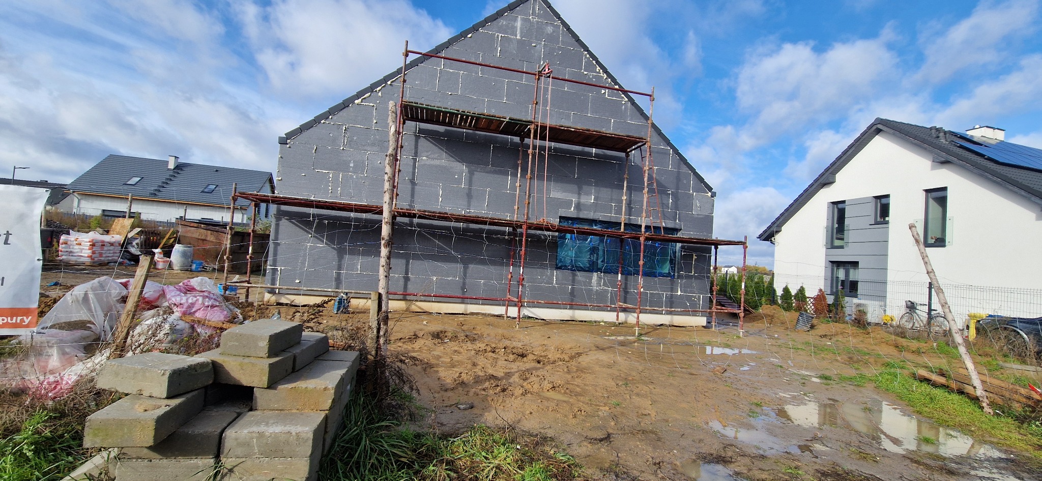 Dom w trakcie ocieplania elewacji, widoczne rusztowania i styropian. Na pierwszym planie stos betonowych bloczków. Błotniste podłoże po deszczu.