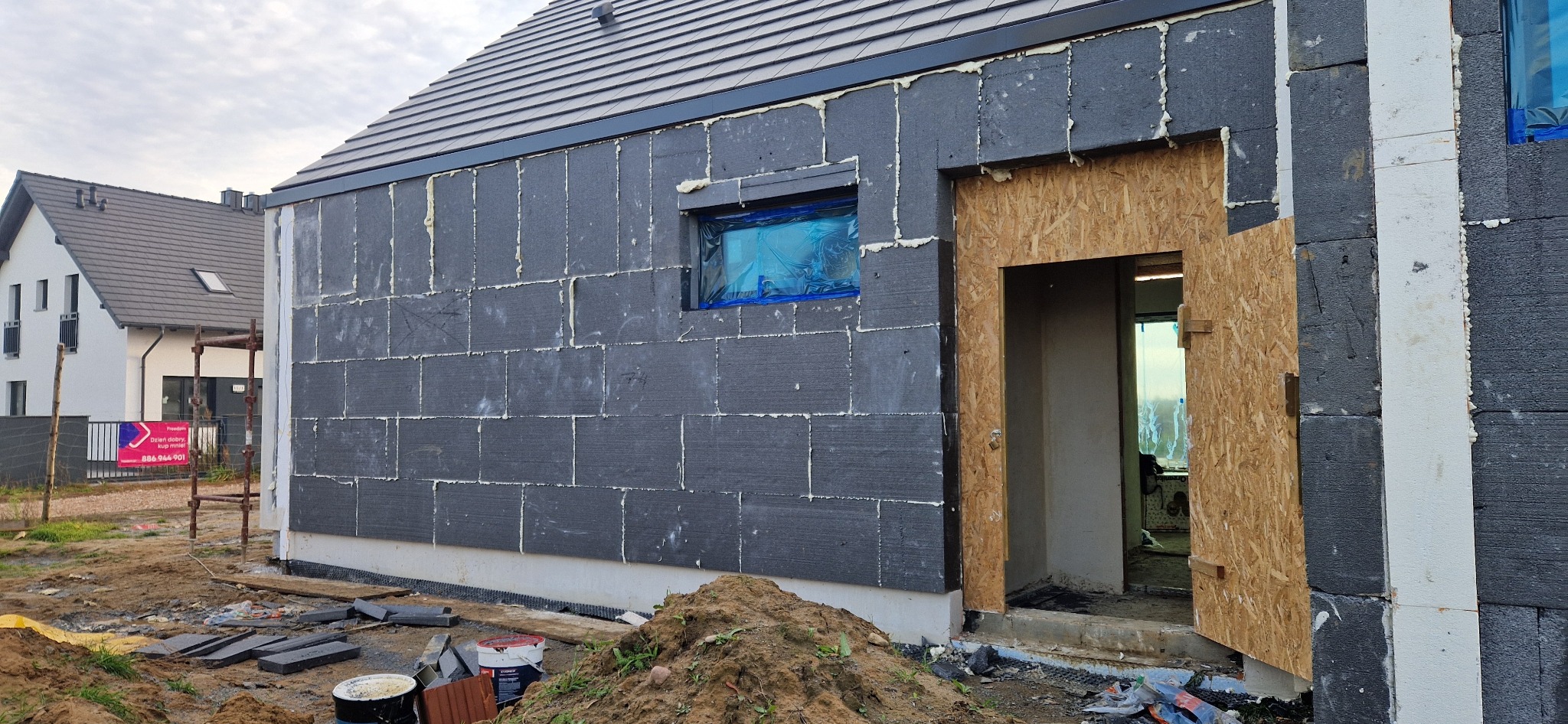 Ściana domu w trakcie ocieplania styropianem grafitowym, widoczne zabezpieczone okno folią i fragment wejścia z płytą OSB. Prace ociepleniowe w toku.