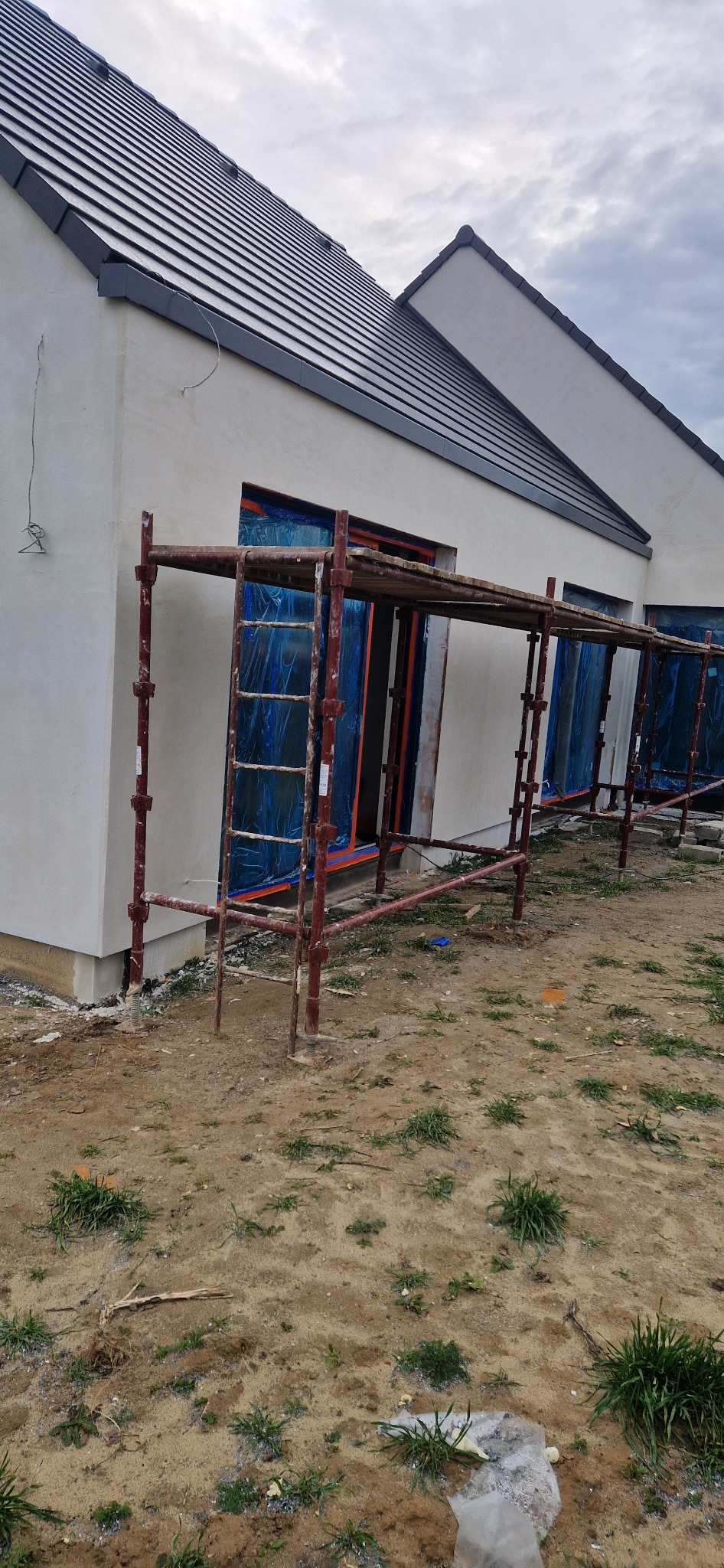 Dom jednorodzinny w trakcie prac elewacyjnych. Widoczne rusztowania, okna zabezpieczone folią, świeżo otynkowana ściana. Ujęcie z poziomu gruntu.