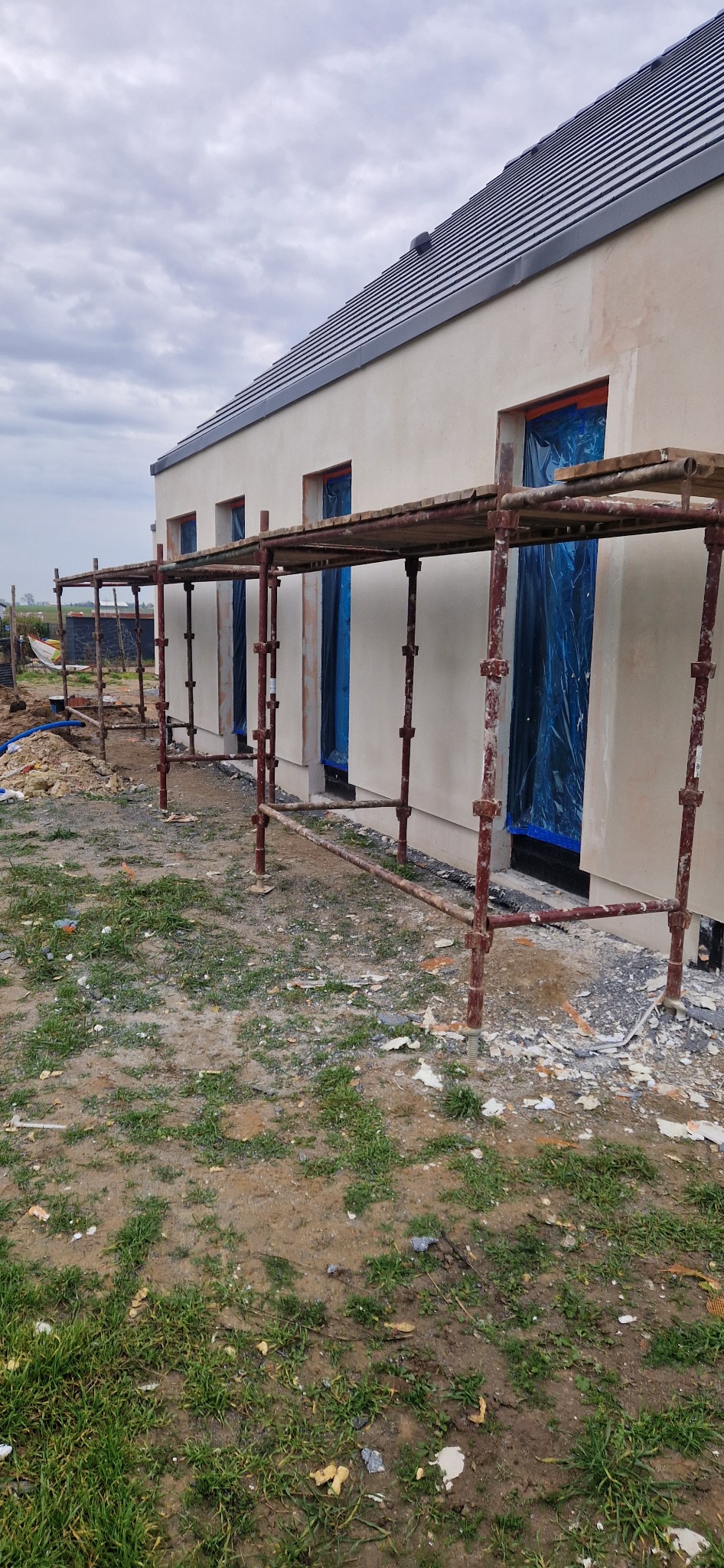 Elewacja domu jednorodzinnego w trakcie prac wykończeniowych. Widoczne rusztowania, okna zabezpieczone folią, świeżo nałożony tynk. Pochmurne niebo w tle.