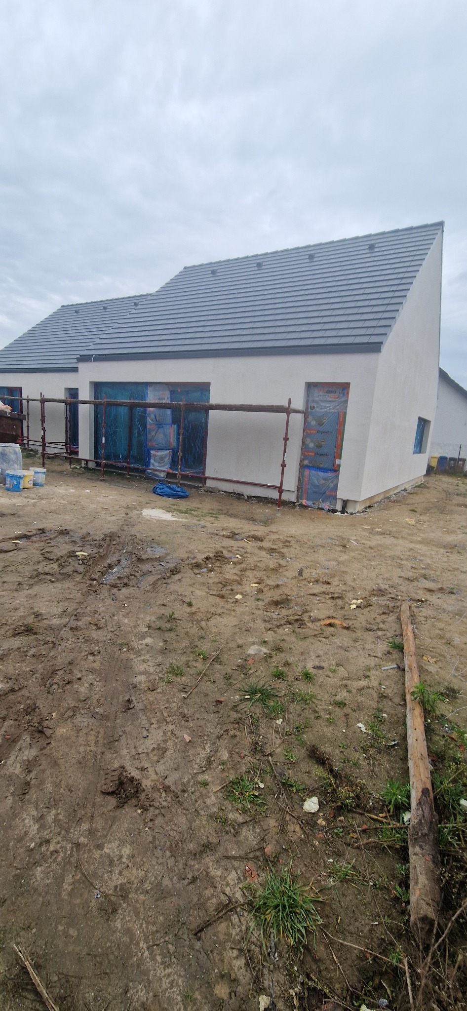 Nowoczesny dom jednorodzinny z szarą dachówką, świeżo otynkowany na biało, z widocznymi rusztowaniami i zabezpieczeniami okiennymi. Teren wokół domu jest jeszcze niezagospodarowany.