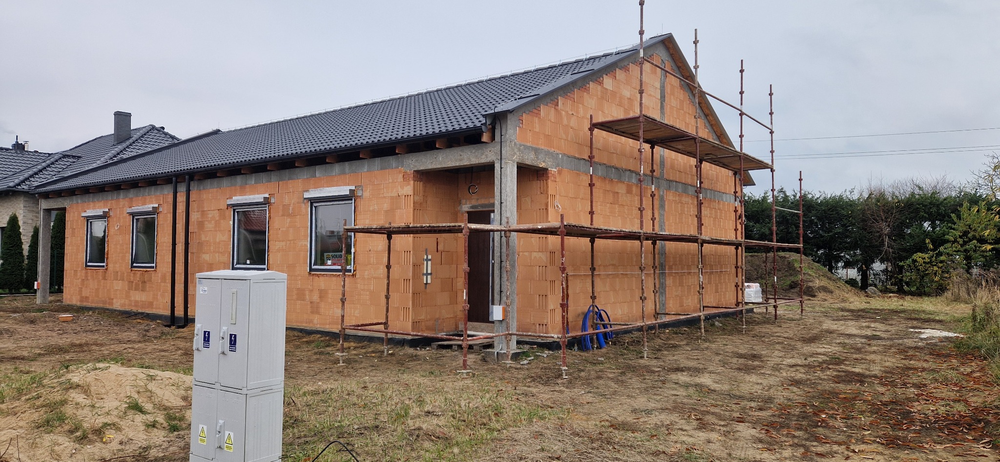 Dom jednorodzinny w trakcie budowy, widoczne ściany z pustaków ceramicznych, okna z roletami zewnętrznymi, rusztowania i dach pokryty dachówką. Stan surowy zamknięty.