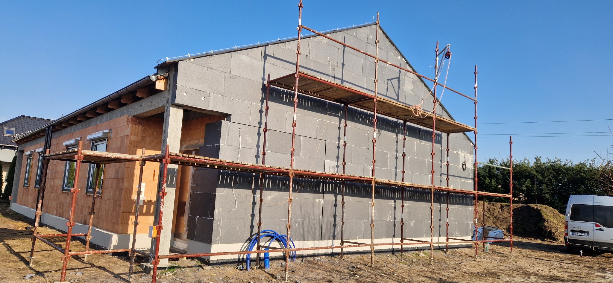 Dom w trakcie ocieplania styropianem, widoczne rusztowania i częściowo otynkowana ściana. Fragment ściany z cegły. Błękitne niebo w tle.