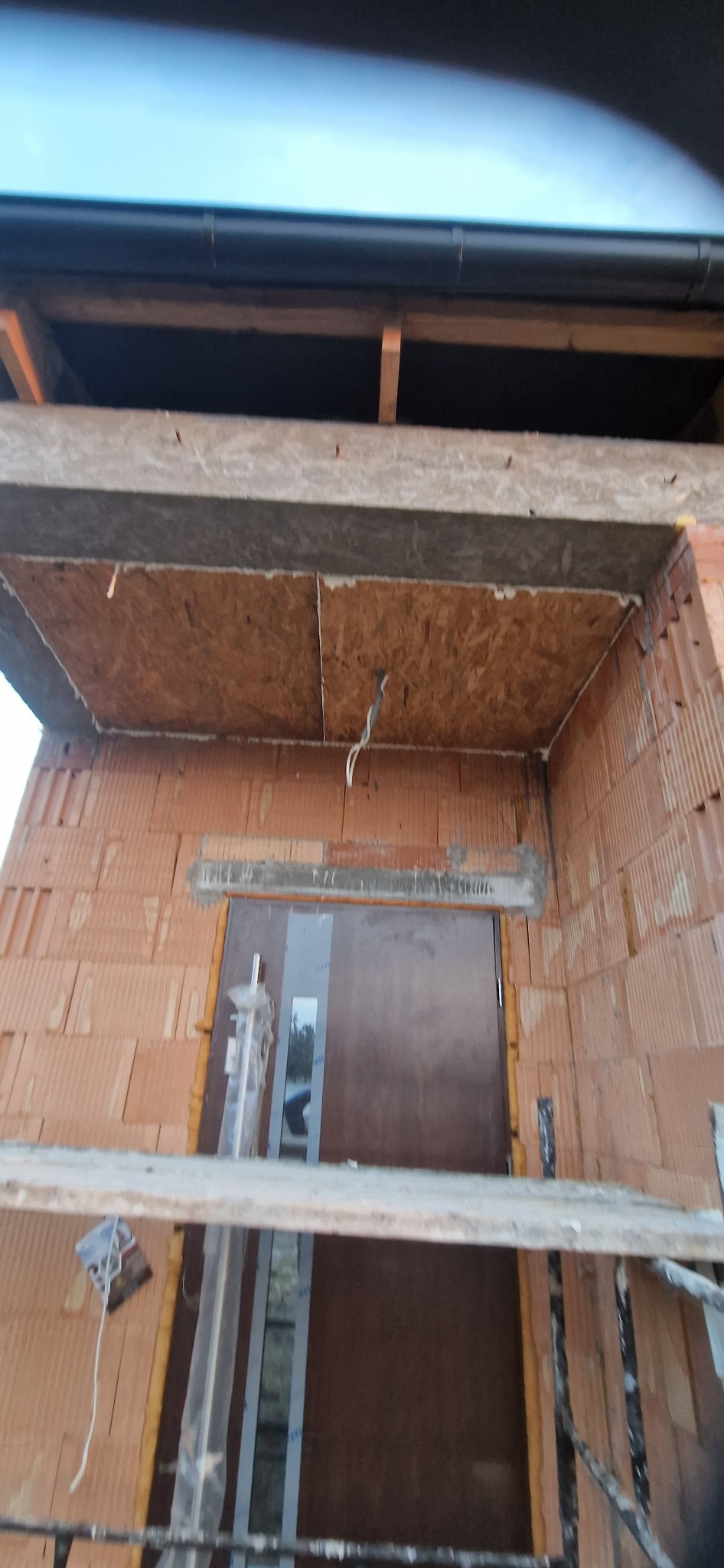 Surowy stan elewacji z pustaków ceramicznych nad brązowymi drzwiami wejściowymi, widoczne elementy konstrukcyjne i instalacja elektryczna, etap budowy.