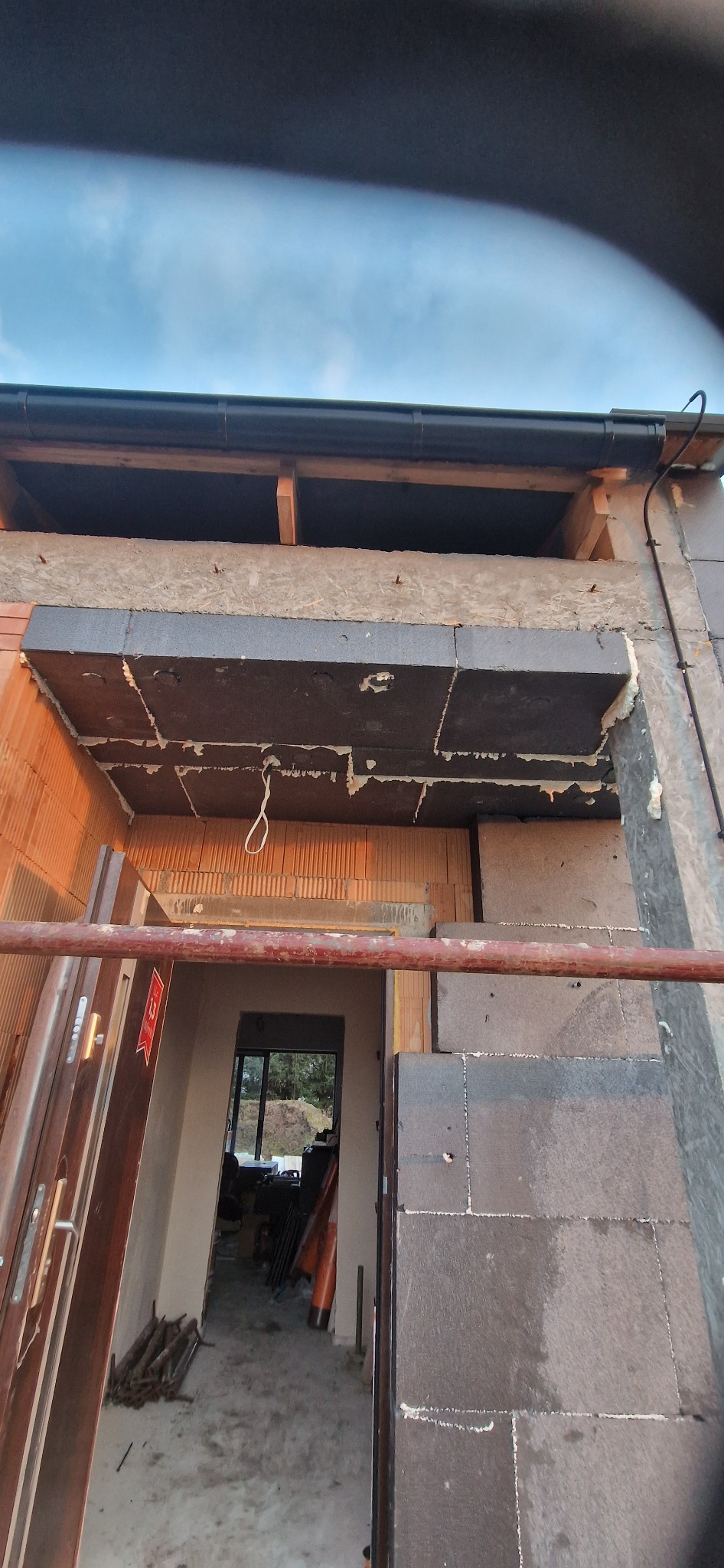 Montaż izolacji termicznej na elewacji budynku z widocznymi elementami rusztowania i fragmentem wnętrza. Użyto czarnego styropianu i pianki montażowej.