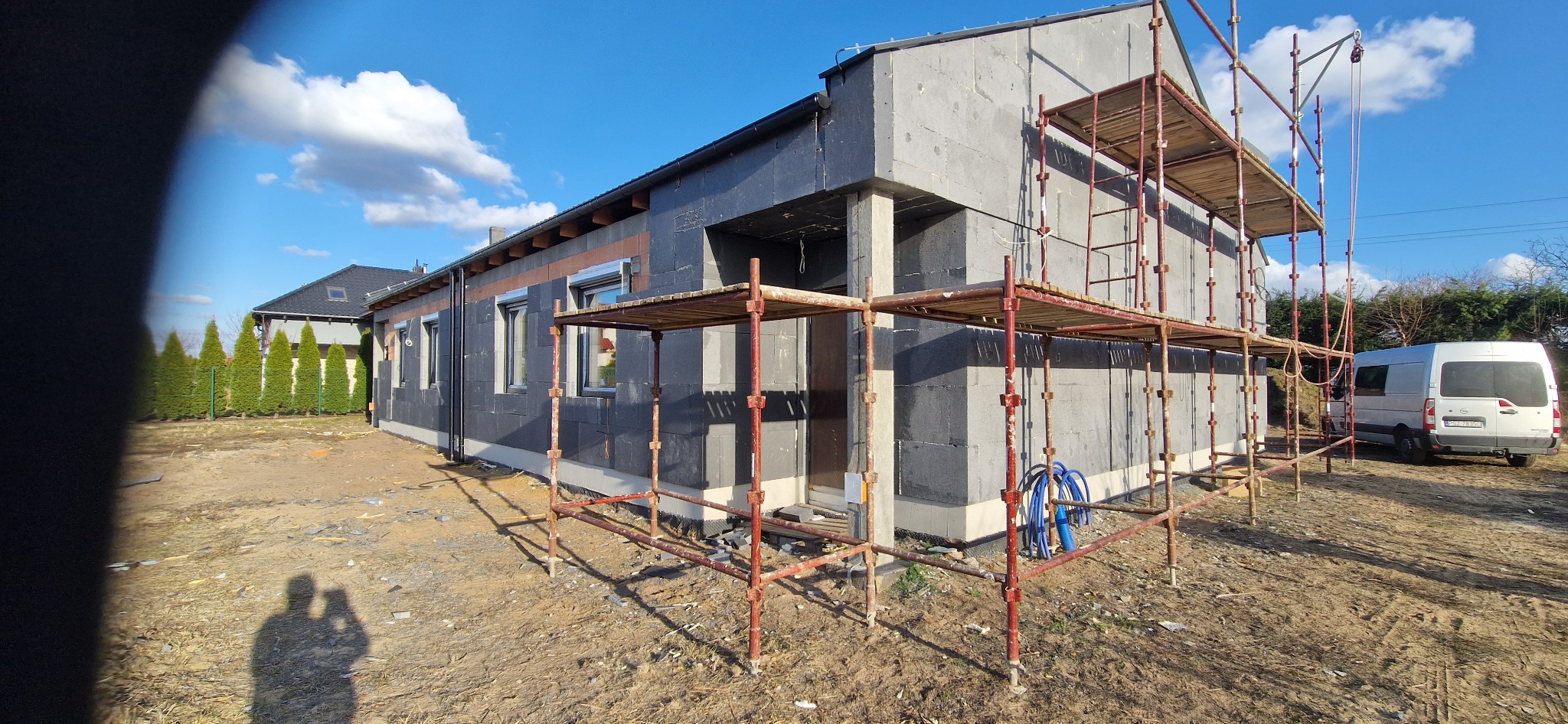 Dom jednorodzinny w trakcie ocieplania, widoczne rusztowania i czarny styropian. Częściowo ocieplona elewacja, błękitne niebo w tle. Na pierwszym planie cień fotografa.