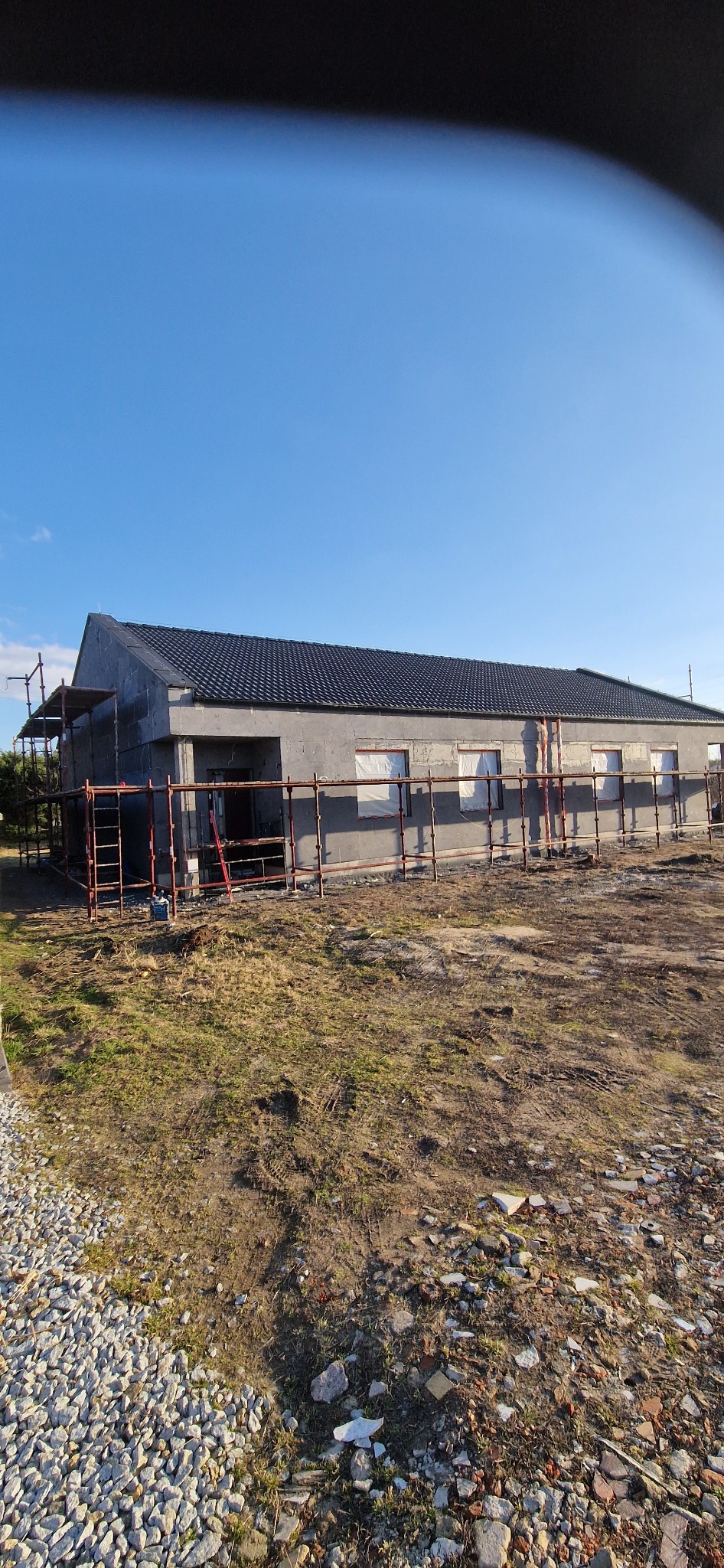 Dom w trakcie ocieplania, widoczne rusztowania i styropian na elewacji. Okna zabezpieczone folią. Dach pokryty ciemną dachówką. Teren wokół budynku niezagospodarowany.