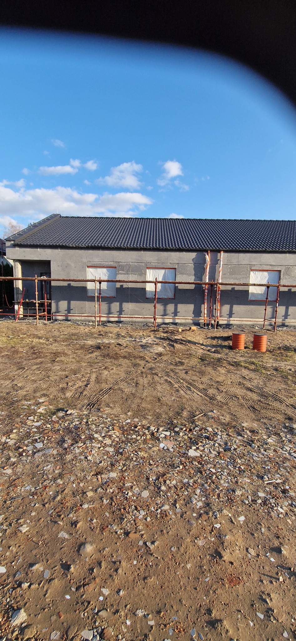 Budynek w trakcie ocieplania, widoczne rusztowania i zabezpieczone okna. Szara elewacja i ciemny dach na tle błękitnego nieba z chmurami. Teren wokół budynku nierówny.
