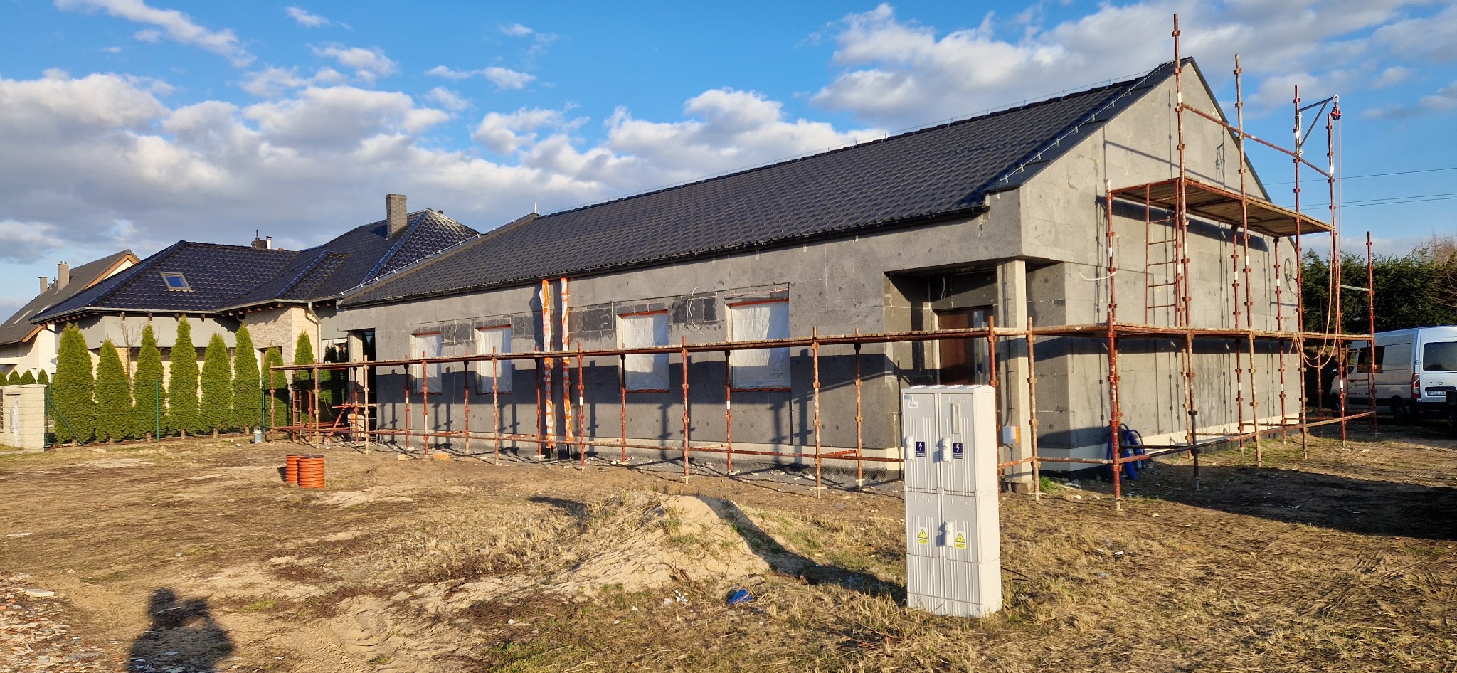 Dom w trakcie ocieplania, widoczne rusztowania i styropian na elewacji. Okna zabezpieczone folią. Na pierwszym planie skrzynka elektryczna.