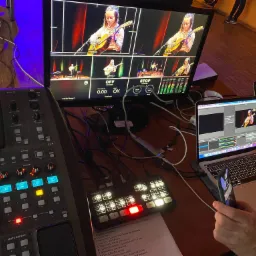 Stanowisko reżyserskie podczas transmisji na żywo z młodą gitarzystką na scenie, widoczne monitory z podglądem kamer, mikser audio-wideo, laptopy z oprogramowaniem do streamingu i kartka z listą...