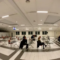 Sala konferencyjna z ustawionymi stolikami i krzesłami, telebimy z wizerunkiem prelegenta, kilka osób w maseczkach siedzi przy stolikach, nagłośnienie na scenie.