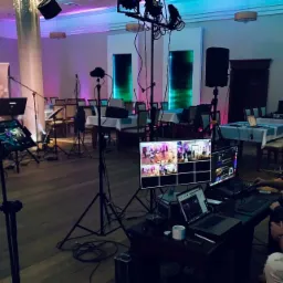 Sala bankietowa przygotowana do imprezy z rozstawionym sprzętem nagłośnieniowym, oświetleniowym i stanowiskiem do miksowania audio/wideo, widoczne monitory z podglądem kamer i laptop...
