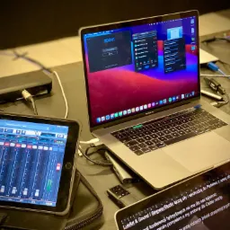 Stanowisko pracy z laptopem MacBook Pro z otwartym Zoomem, tabletem z aplikacją miksera audio i drugim tabletem wyświetlającym tekst o konferencjach hybrydowych, kable i adaptery na stole...