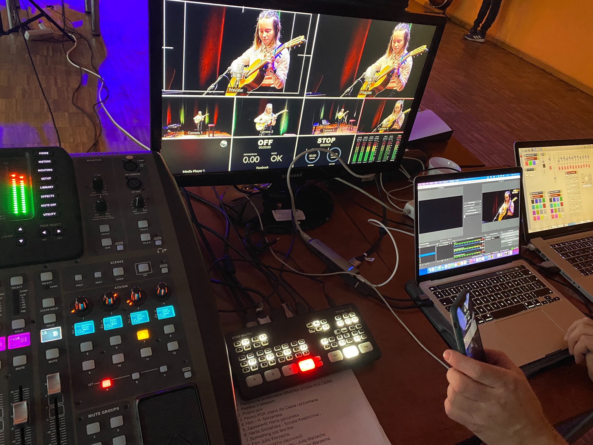 Stanowisko reżyserskie podczas transmisji na żywo z młodą gitarzystką na scenie, widoczne monitory z podglądem kamer, mikser audio-wideo, laptopy z oprogramowaniem do streamingu i kartka z listą...