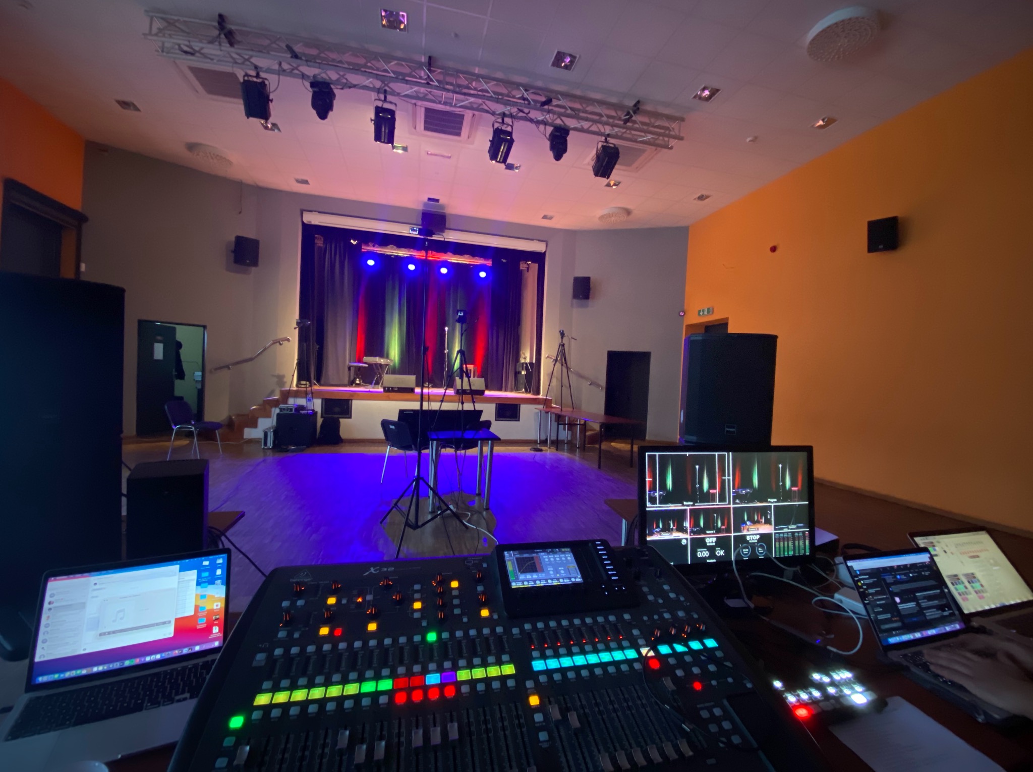 Widok zza konsolety dźwiękowej na scenę oświetloną kolorowymi światłami, z widocznymi laptopami i ekranami monitorującymi poziom dźwięku.