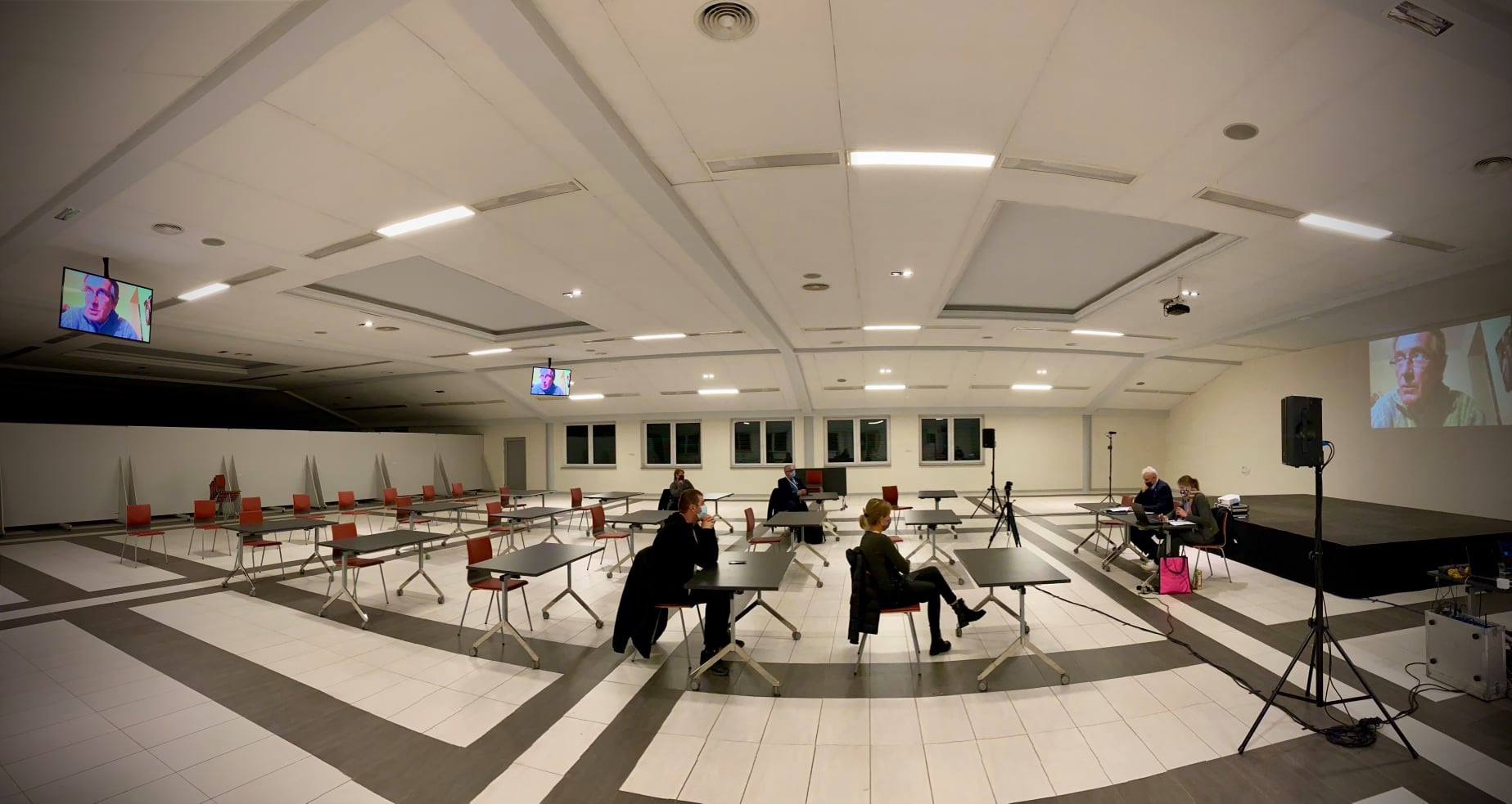 Sala konferencyjna z ustawionymi stolikami i krzesłami, telebimy z wizerunkiem prelegenta, kilka osób w maseczkach siedzi przy stolikach, nagłośnienie na scenie.