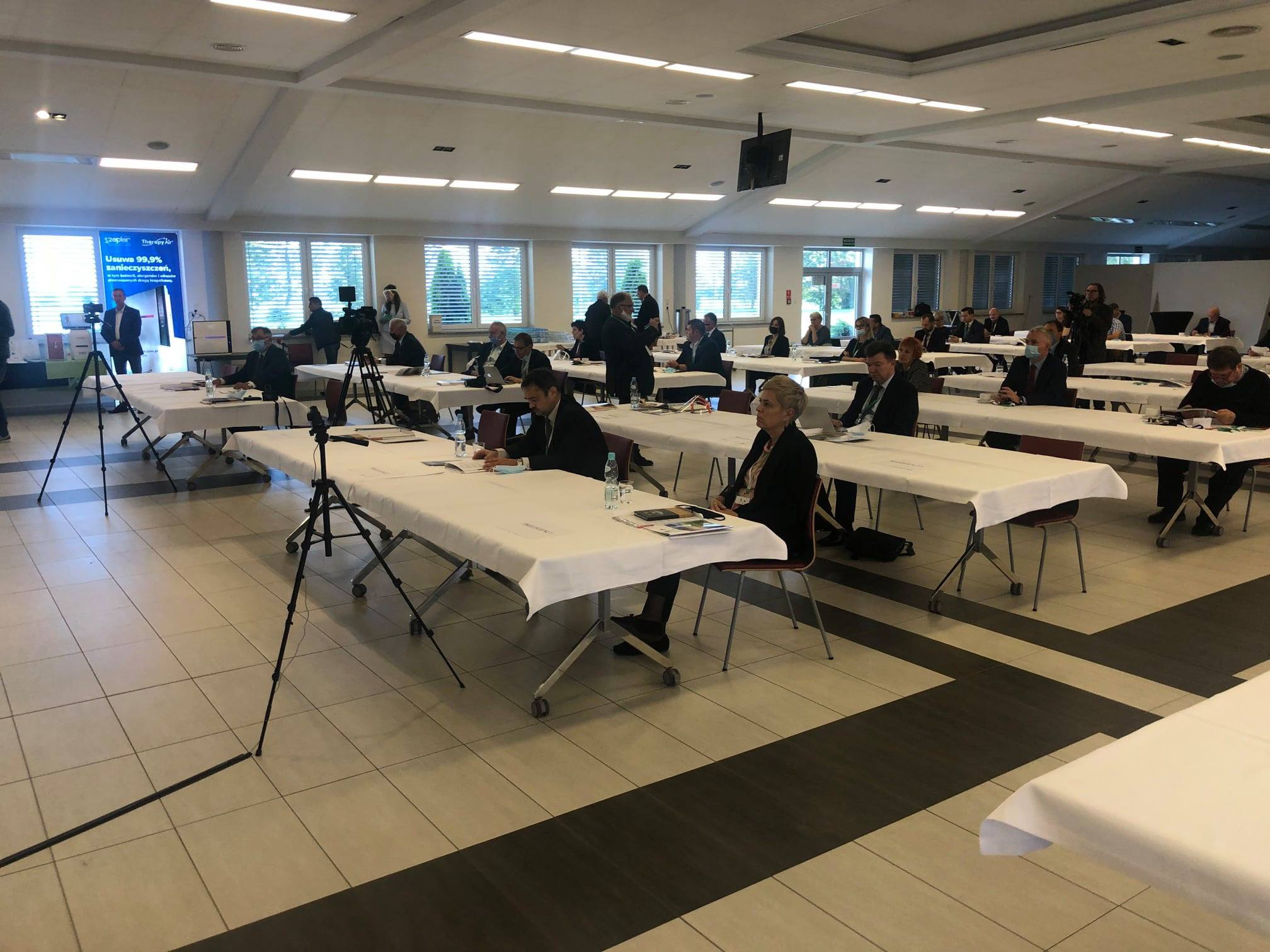 Sala konferencyjna z uczestnikami siedzącymi przy stołach z białymi obrusami, oświetlenie sufitowe, kamery na statywach dokumentujące wydarzenie, osoby w maskach.