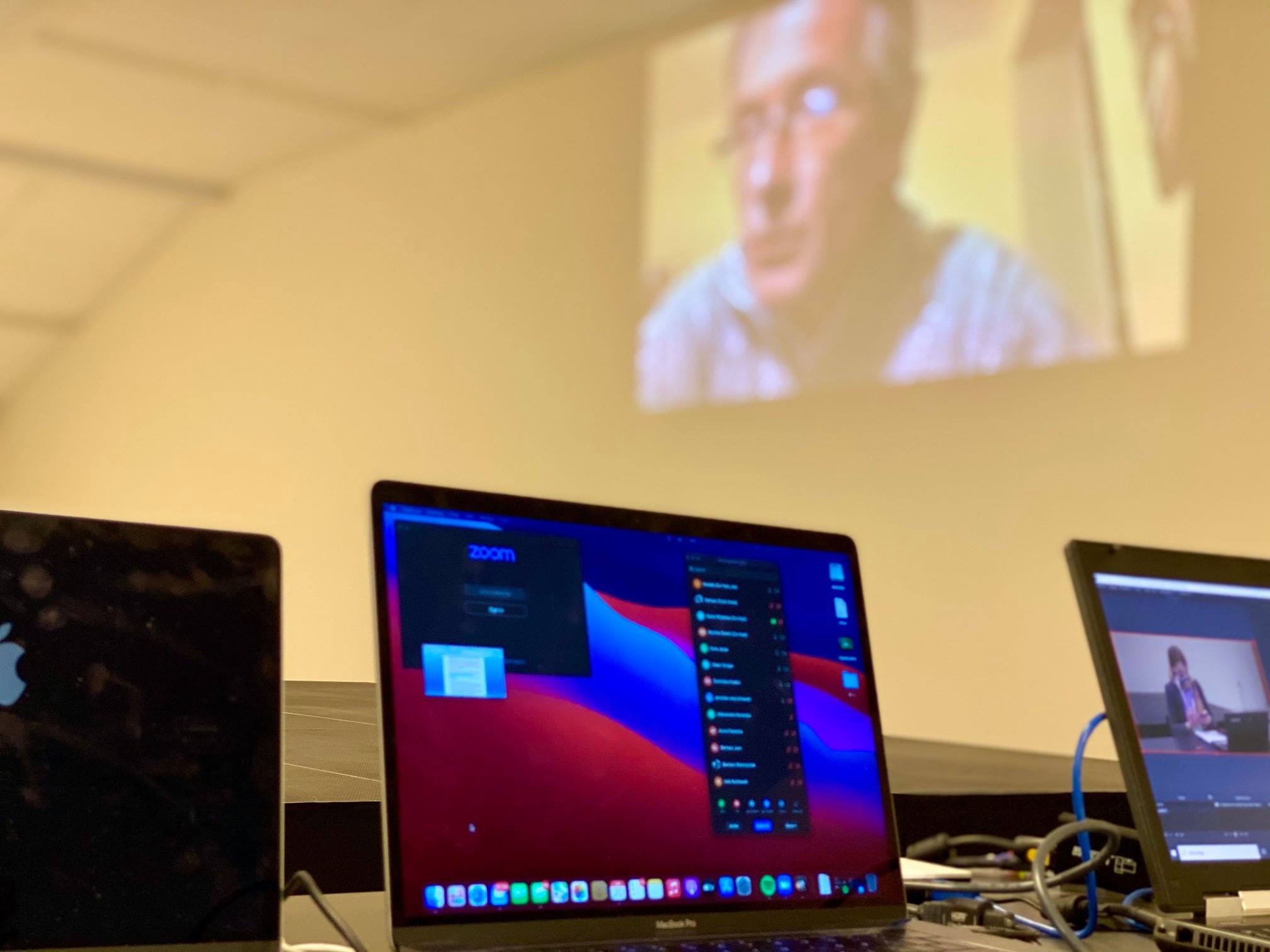 Trzy laptopy na stole, środkowy z otwartym programem Zoom, na ścianie wyświetlany obraz z wideokonferencji.