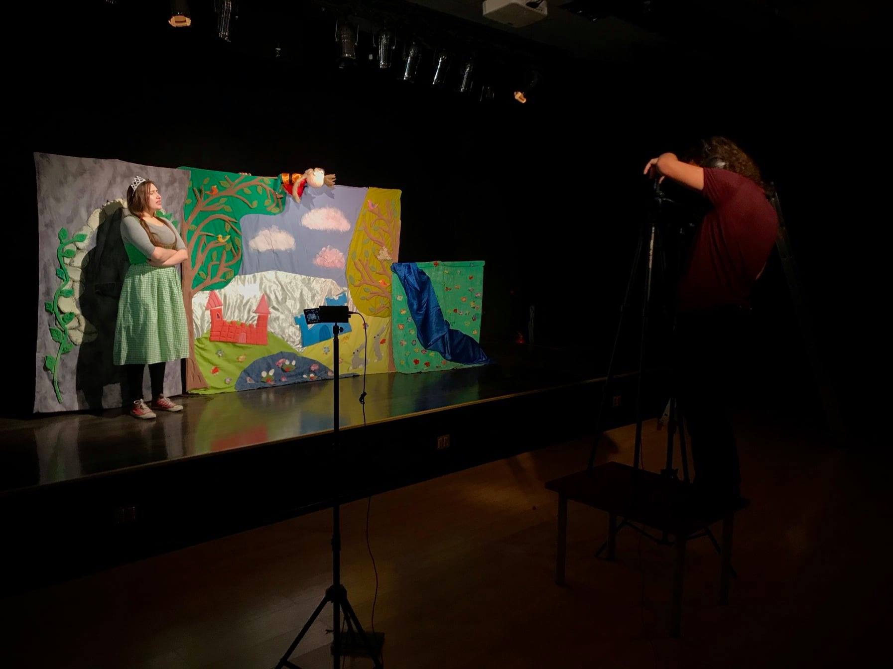 Scena teatralna z aktorką w kostiumie księżniczki na tle ręcznie robionej scenografii, filmowana przez kamerzystę z boku sceny.